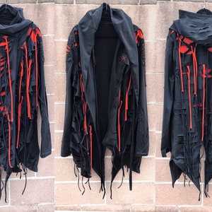 May include: Black hooded jacket with red accents and distressed detailing. The jacket has long sleeves and a long, asymmetrical hemline. The fabric is frayed and has a ripped look.