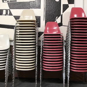 May include: A collection of stacked chairs in various colors, including cream and red. The chairs have a minimalist design with metal legs and are arranged in neat stacks, showcasing a modern aesthetic.