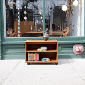 May include: A small, two-shelf wooden bookcase on wheels, filled with books and decorative items. The bookcase is a warm brown color and sits in front of a green storefront. A vase of flowers sits on top.