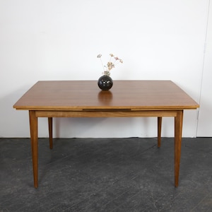 May include: A rectangular wooden dining table with a warm brown finish. The table has four tapered legs and a dark vase with dried flowers on top. The table is approximately 60 inches long.
