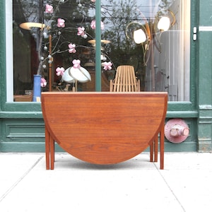 May include: A mid-century modern, half-circle wooden table with four slender legs. The table is a warm, reddish-brown color and appears to be made of solid wood. The table is set up on a sidewalk in front of a storefront.