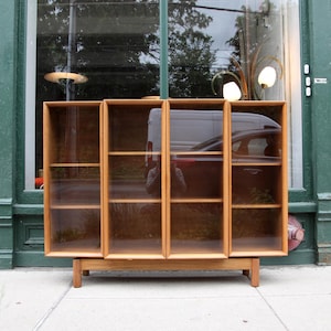 May include: A light brown wooden bookcase featuring four vertical glass-fronted sections. The unit is supported by a rectangular base with four short legs and contains multiple horizontal shelves visible through the clear glass panels.