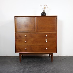 May include: A mid-century modern wooden dresser with a hutch. The dresser has three drawers with white knobs and a top cabinet with two doors. A small black vase with dried flowers sits on top. The dresser is made of a warm-toned wood.