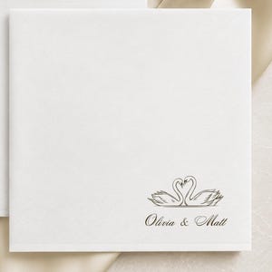 May include: Two square white paper napkins featuring a black line drawing of two swans forming a heart shape with their necks. Below the swan illustration, the text reads Olivia & Matt in a cursive font. The napkins are displayed on a champagne fabric.