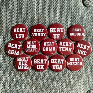 May include: A collection of maroon buttons with white text. Each button has the word "BEAT" above a university's abbreviation, including LSU, Vandy, UF, Auburn, A&M, Miss State, Bama, Tenn, USC, Ole Miss, UK, UGA, and Mizzou.