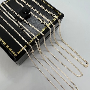 May include: Multiple silver-coloured mariner link chain necklaces are arranged on a black jewellery box with gold trim. The chains, of different lengths, are displayed on a white surface. The chains are made of a shiny metal.
