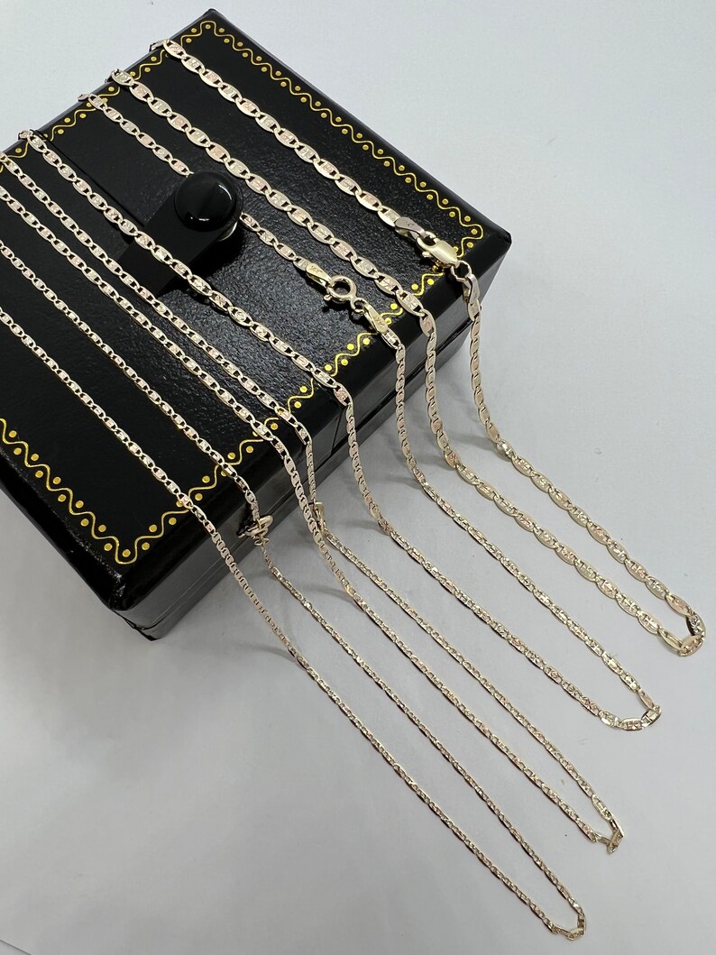 May include: Multiple silver-coloured mariner link chain necklaces are arranged on a black jewellery box with gold trim. The chains, of different lengths, are displayed on a white surface. The chains are made of a shiny metal.