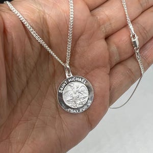 May include: A silver Saint Michael pendant necklace. The round pendant features an image of Saint Michael and the words "Saint Michael Pray For Us". The chain is a delicate curb link style.