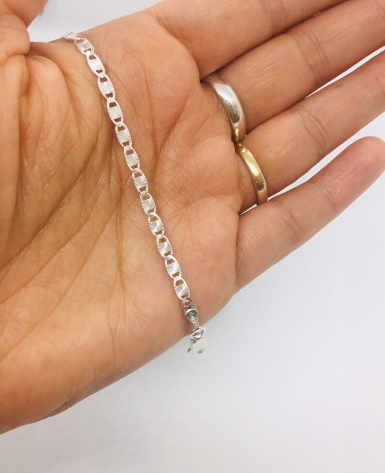 May include: A silver chain bracelet with a delicate, patterned design. The chain is made of small, oval-shaped links.