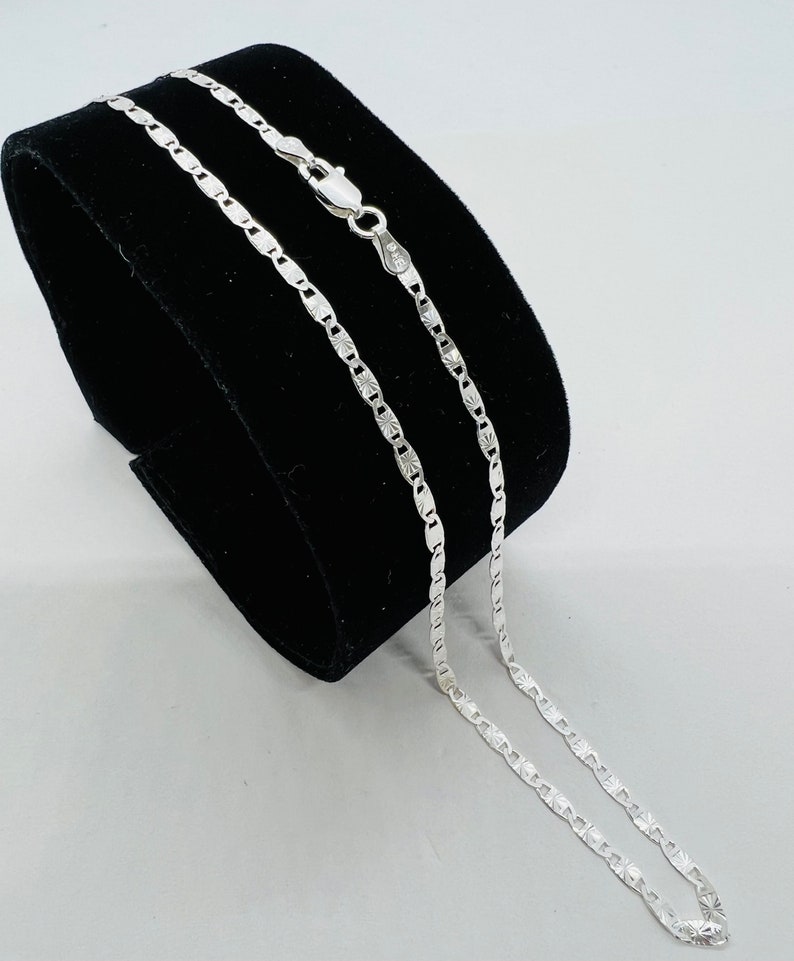 Puede incluir: Un collar de cadena de plata con un dise&ntilde;o de cadena de caja. La cadena est&aacute; en un expositor de terciopelo negro.
