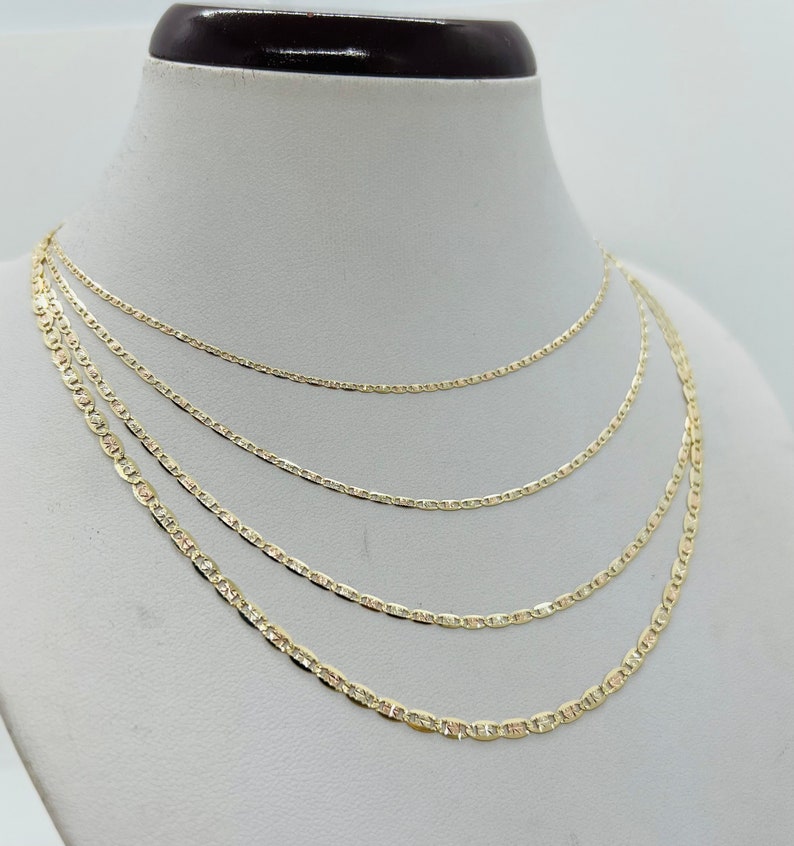 May include: Four gold-coloured chain necklaces of varying lengths displayed on a white mannequin bust. The chains have a flat, mariner link design. The necklaces are arranged to show different lengths and styles.