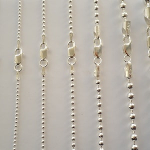 May include: Seven silver chains with ball links. The chains are all the same length and are hanging from a white background.