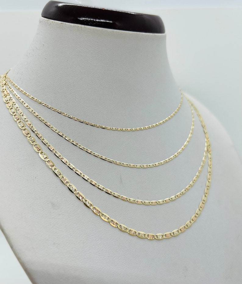 May include: Four gold-coloured mariner chain necklaces of varying thicknesses displayed on a white mannequin bust. The chains have a flat, oval link design, and are presented in a layered arrangement, showcasing the different sizes.
