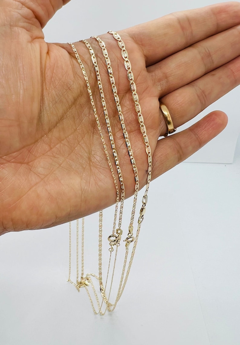 May include: A selection of gold-coloured chain necklaces in various styles and thicknesses. The chains are held against a light background, highlighting their different designs and clasps. The necklaces appear to be made of a shiny, metallic material.