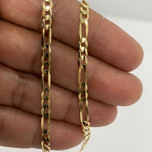 May include: A gold chain necklace with a figaro link pattern. The chain is shown on a hand, but the hand is not visible in the image.