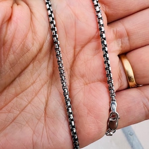 May include: A silver-colored box chain necklace with a lobster clasp. The chain is held in a hand, and the clasp is marked "Italy". The chain is made of small, square links.