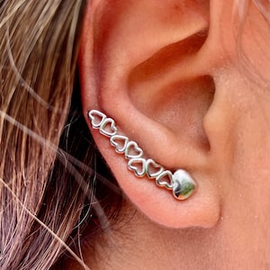 May include: Silver ear cuff with a row of connected heart shapes and a single heart charm at the end.
