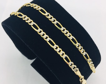 May include: Two gold chains, one with a yellow gold finish and the other with a white gold finish, are displayed on a black velvet stand. The chains are both figaro style.