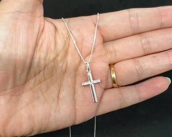 May include: A silver cross pendant necklace. The cross is a simple, polished design, suspended from a delicate chain. The necklace is displayed against a dark background.