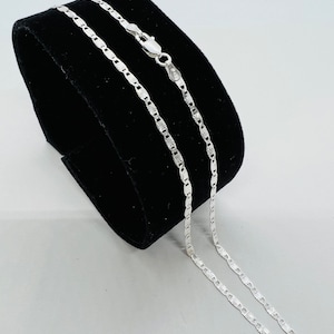 May include: A silver chain necklace with a delicate, patterned design. The chain is displayed on a black velvet background.