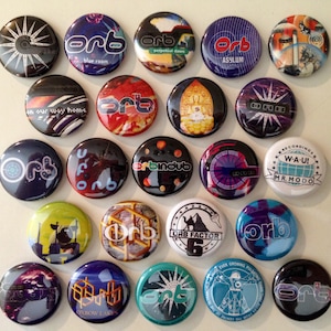 May include: A collection of 20 round buttons with the word "orb" printed on them. Each button has a different design, including a starburst, a cityscape, a Buddha, and a geometric pattern. The buttons are all in various shades of blue, green, red, and yellow.