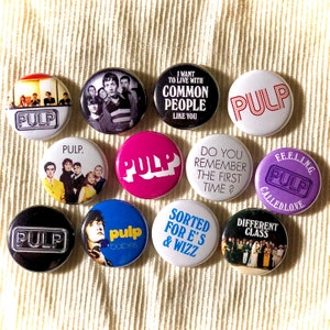 Op de afbeelding: Een verzameling van 10 ronde, kleurrijke knoppen met het woord "PULP" erop gedrukt. De knoppen tonen verschillende albumhoezen en songtitels van de band Pulp.