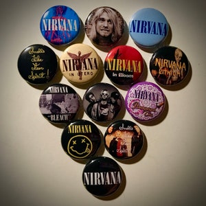 May include: A collection of 11 round Nirvana band pins. The pins feature various album covers, band logos, and lyrics. The pins are black, white, blue, red, purple, and yellow.