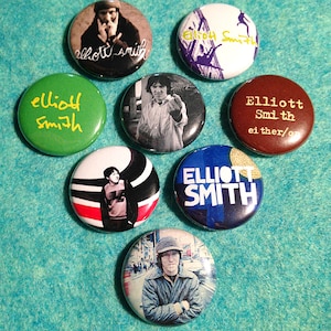 May include: Eight different button pins featuring a portrait of musician Elliott Smith. The pins are round and feature various images and text, including a portrait of Elliott Smith, a cityscape, and the text "Elliott Smith".