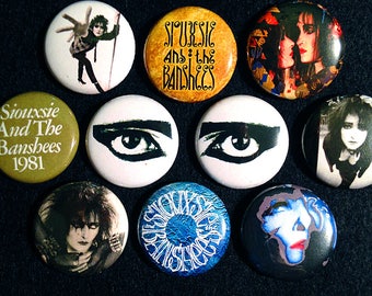 Puede incluir: Nueve botones redondos con pasador que muestran imágenes de la banda Siouxsie and the Banshees. Los botones presentan una variedad de imágenes, incluyendo un retrato de Siouxsie Sioux, un primer plano de un ojo y el nombre de la banda en una fuente estilizada.