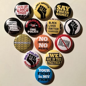 May include: Twelve round button badges with various messages and images related to the Black Lives Matter movement. The badges feature phrases like "Black Lives Matter", "Say Their Names", "No Justice No Peace", and "We Are In This Together". Some badges also feature images of raised fists and other symbols of protest.