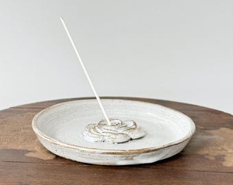 May include: A white ceramic incense holder with a rose design in the center. The holder is a shallow dish with a slightly raised edge. A white incense stick is inserted into the rose. The holder sits on a wooden surface.