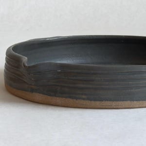 May include: A shallow, circular ceramic dish with a dark charcoal gray glazed interior and exterior rim. The exterior features horizontal ridges and a small indentation along the edge, while the base remains unglazed in a natural tan clay color.