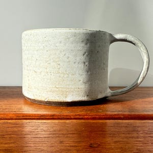 May include: A handmade ceramic mug with a textured, off-white exterior and a dark brown base. The mug has a rounded handle and sits on a wooden surface. This stoneware mug is perfect for coffee or tea.