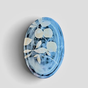 Puede incluir: Un plato ovalado de cerámica con un esmalte jaspeado en azul y blanco. La superficie está decorada con una ilustración botánica clara de una rama con frutos redondos y hojas, creando un contraste sobre el fondo azul oscuro.