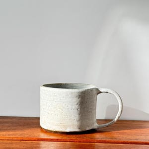 May include: A handmade, off-white ceramic mug with a rounded handle. The mug has a textured surface and a dark brown base. The mug is sitting on a wooden surface.