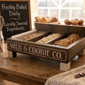 Może przedstawiać: Ciemnobrązowa drewniana taca ekspozycyjna z trzema przegródkami, z białym napisem MILK & COOKIE CO. z przodu. Taca posiada cztery jasne, kwadratowe nóżki i jest wypełniona różnymi okrągłymi ciasteczkami ułożonymi w rzędach.