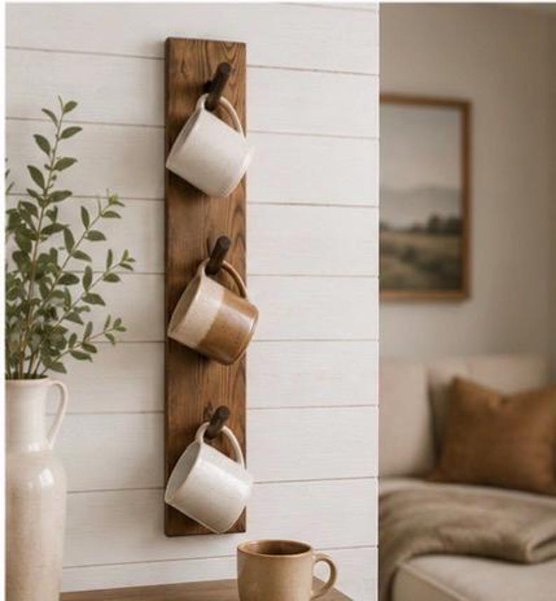 Może przedstawiać: Pionowy drewniany wieszak ścienny z trzema ciemnobrązowymi kołkami, na kt&oacute;rych zawieszono ceramiczne kubki. G&oacute;rny i dolny kubek są w kolorze złamanej bieli, a środkowy jest dwukolorowy, w odcieniach beżu i kremu. Dodatkowy beżowy kubek stoi na blacie.