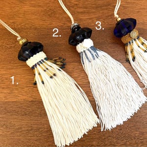 May include: Three decorative tassels with white fringe and beaded accents. Each tassel features a different top design: black, black and white, and blue. The tassels are displayed on a wooden surface.
