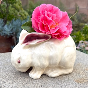 May include: A white ceramic bunny figurine with pink inner ears and red eyes, holding a vibrant pink flower. The bunny is placed on a gray surface, with greenery in the background.