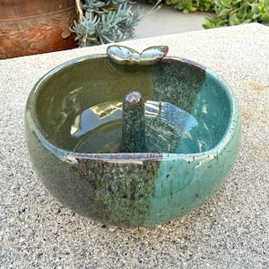 May include: A handcrafted ceramic bowl with a unique design. The bowl features a two-tone glaze in shades of green and teal, with a central post and a leaf-shaped accent. The bowl is likely used for decorative purposes.
