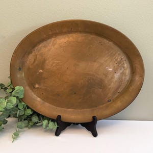 May include: A large, oval copper tray with a warm, metallic finish. The tray is displayed on a black stand, with green foliage to the left. The tray's surface shows slight variations in color, adding to its vintage appeal.
