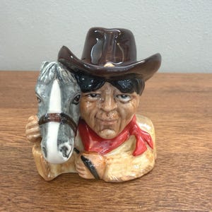 May include: A ceramic figurine of a cowboy with a brown hat and red scarf, holding a horse's head. The cowboy has a tan shirt and the horse is gray and white. The figurine is on a wooden surface.