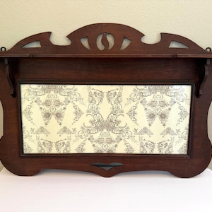 May include: Dark brown wooden wall shelf with a decorative frame. The shelf has a rectangular center with a cream-colored background and a gray floral pattern. The top of the shelf has a decorative cutout design.