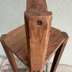 May include: A vintage wooden plant stand with a multi-tiered design. The stand has a central, vertical support with two hexagonal shelves. The wood has a warm, reddish-brown tone, showing the natural grain and texture of the material.