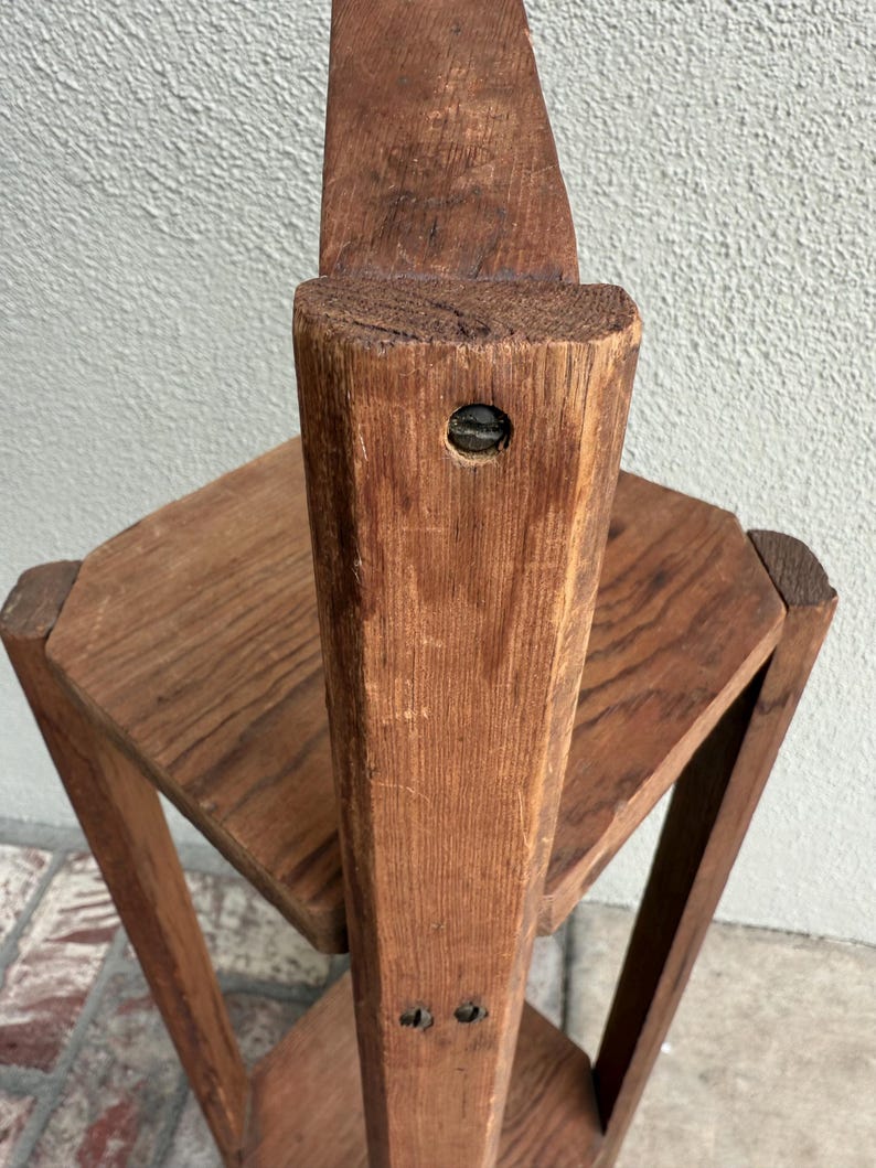 May include: A vintage wooden plant stand with a multi-tiered design. The stand has a central, vertical support with two hexagonal shelves. The wood has a warm, reddish-brown tone, showing the natural grain and texture of the material.
