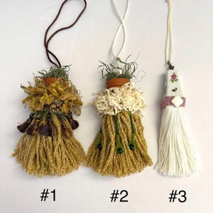 May include: Three decorative tassels with unique designs. The first two feature small terracotta pots with greenery, surrounded by textured yarn in shades of gold and cream. The third tassel has a porcelain top with floral details and a white tassel.