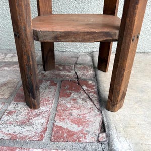 May include: A small, rustic wooden stool with a square seat and four angled legs. The wood has a warm, brown tone with visible grain and knots, suggesting a handmade or antique piece. The stool is sturdy and simple in design.