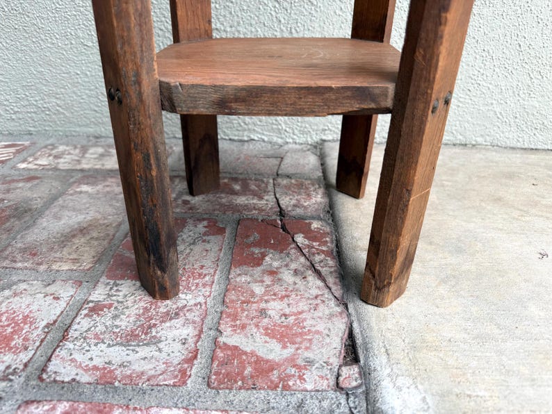 May include: A small, rustic wooden stool with a square seat and four angled legs. The wood has a warm, brown tone with visible grain and knots, suggesting a handmade or antique piece. The stool is sturdy and simple in design.
