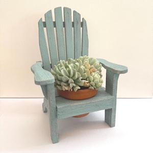 May include: A miniature Adirondack chair painted in a distressed teal blue, holding a small terracotta pot with a succulent plant. The chair has a slatted back and armrests, evoking a coastal or garden theme.