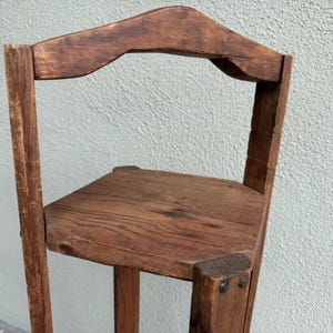 May include: A small, rustic wooden corner shelf with a triangular seat and a curved backrest. The shelf is made of dark brown wood and has visible wood grain. The construction features visible nails and a simple, functional design.